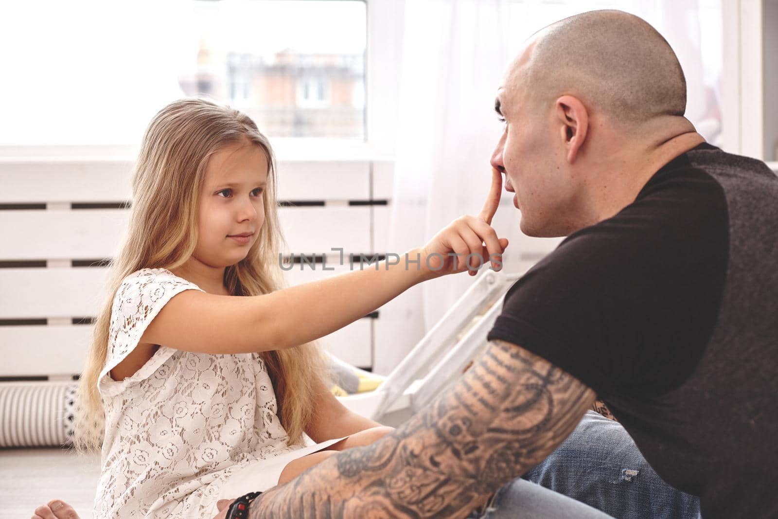 White modern kid's room whith a wooden furniture. Adorable daughter wearing a white dress is touching daddy's nose and looking with tenderness at him. Young daddy with tattoos is hugging her. Friendly family spending their free time together sitting on a pillows.