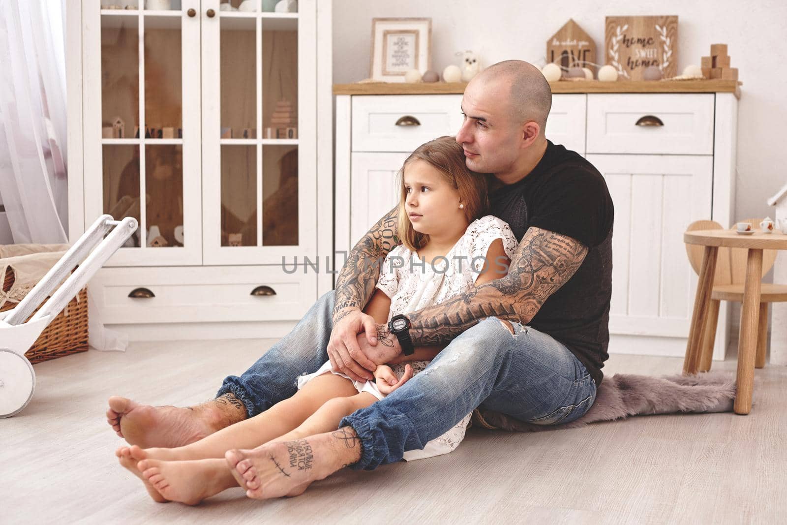 White modern kid's room whith a wooden furniture. Adorable daughter with a long blond hair wearing a white dress. Daddy with tattoos is hugging her and they are looking away. Friendly family spending their free time together sitting on a pillows.