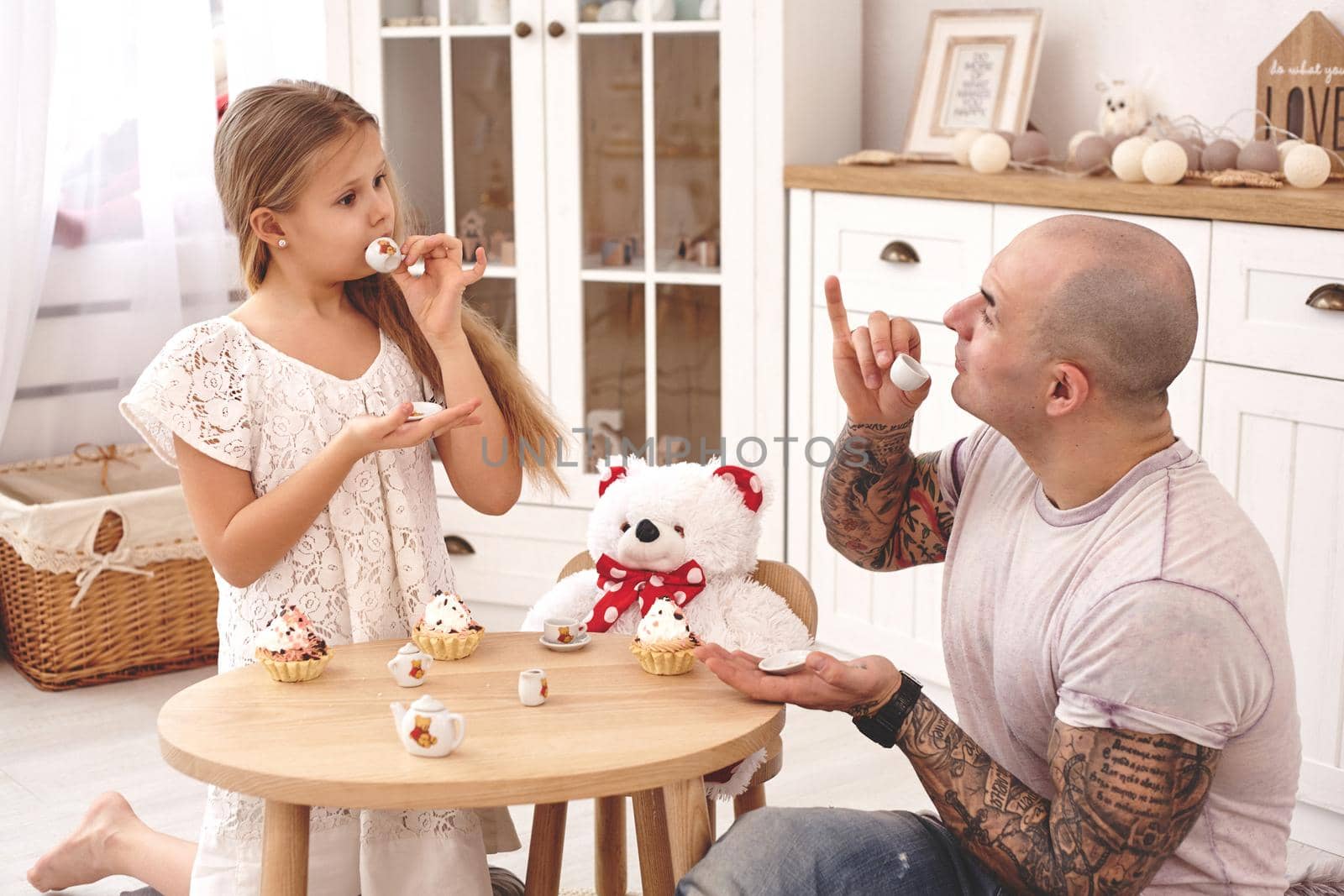 White modern kid's room whith a wooden furniture. Adorable daughter whith a long blond hair wearing a white dress. They are drinking tea with cupcackes from a toy dishes in a modern kid's room whith a wooden furniture. Daddy with tattoos is looking at his daughter and she is looking at him also. Friendly family spending their free time together sitting on a pillows.