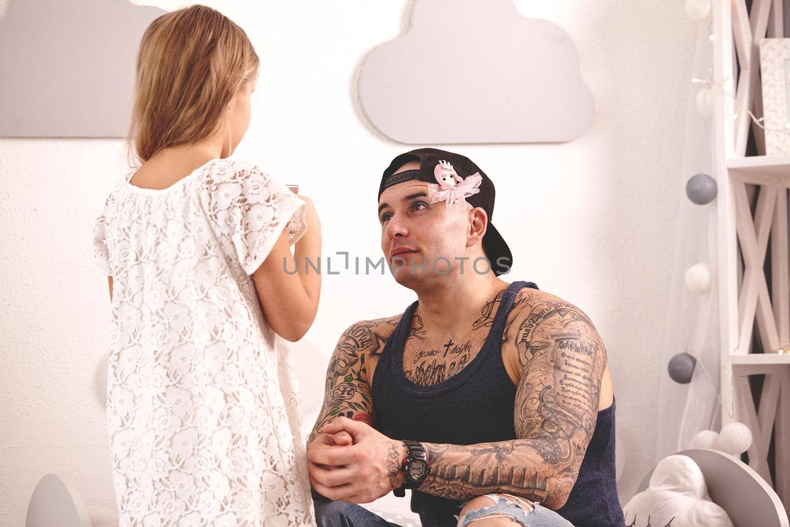 Funny time Tattoed father and his child are playing at home. Cute girl in a white dress is doing makeup to her dad in her bedroom. Daddy is looking at her whith love. Family holiday and togetherness.