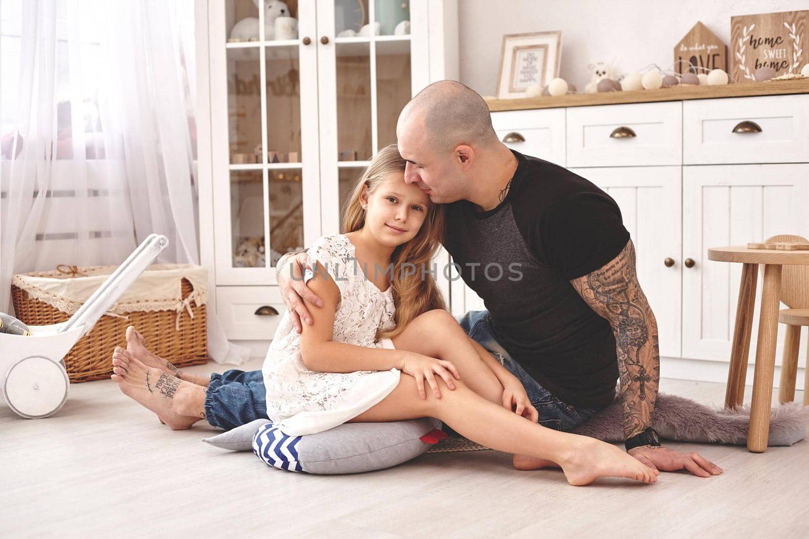 White modern kid's room whith a wooden furniture. Adorable daughter with a blond long hair wearing a white dress. Daddy with tattoos is kissing and hugging her. Friendly family spending their free time together sitting on a pillows.