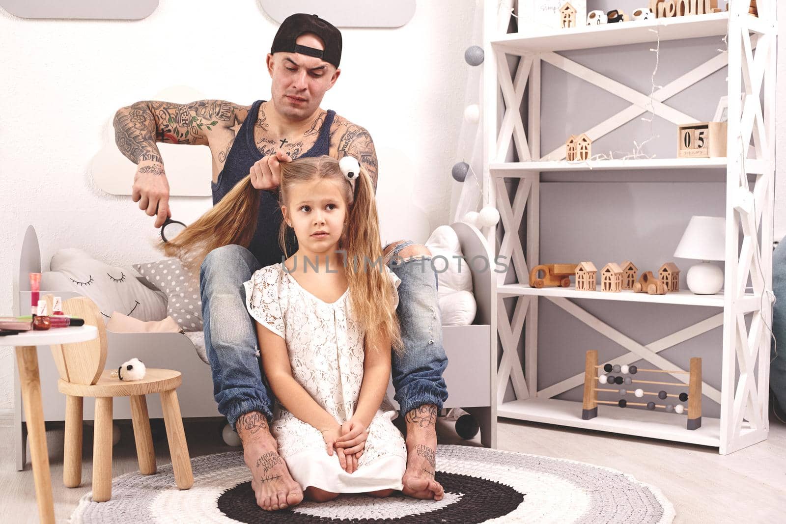 Funny time Attractive father in a cap and his child are playing at home. Dad is doing his daughter's hair while she is sitting on a rug in her bedroom. Family holiday and togetherness.