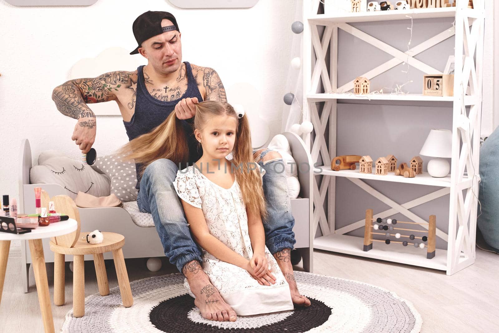 Funny time Tattoed father in a cap and his child are playing at home. Dad is doing his daughter's hair in her bedroom. Family holiday and togetherness by nazarovsergey