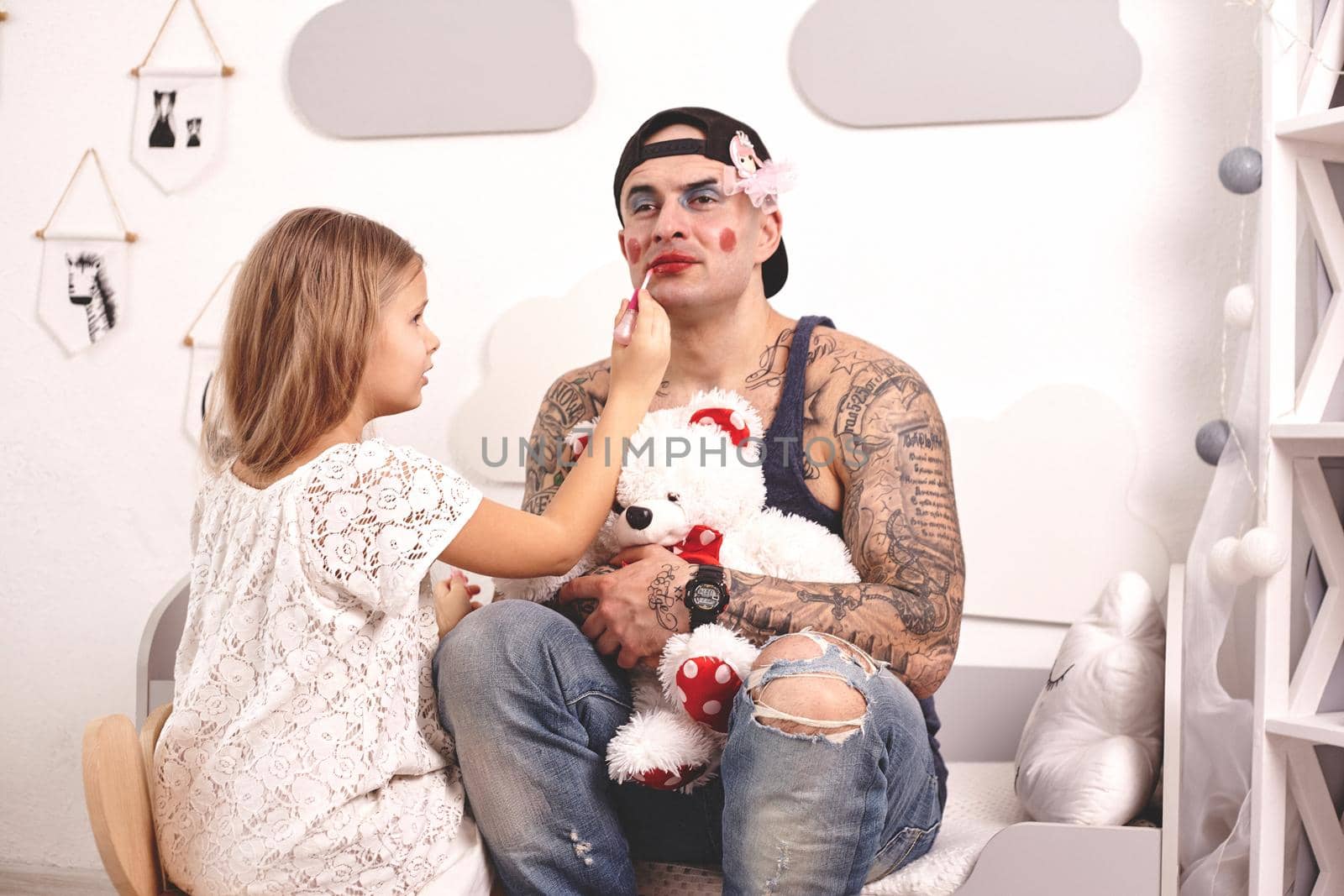 Funny time Tattoed father in a cap and his child are playing at home. Pretty girl in a white dress is putting a lipstick at her dady's lips while he is holding a toy bear, in the bedroom. Family holiday and togetherness.