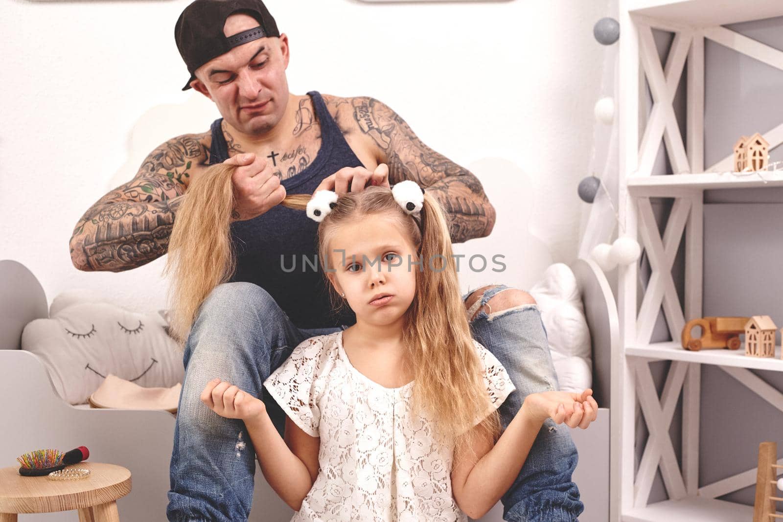 Funny time Tattoed father in a cap and his child are playing at home. Dad is doing his daughter's hair in her bedroom. Family holiday and togetherness.