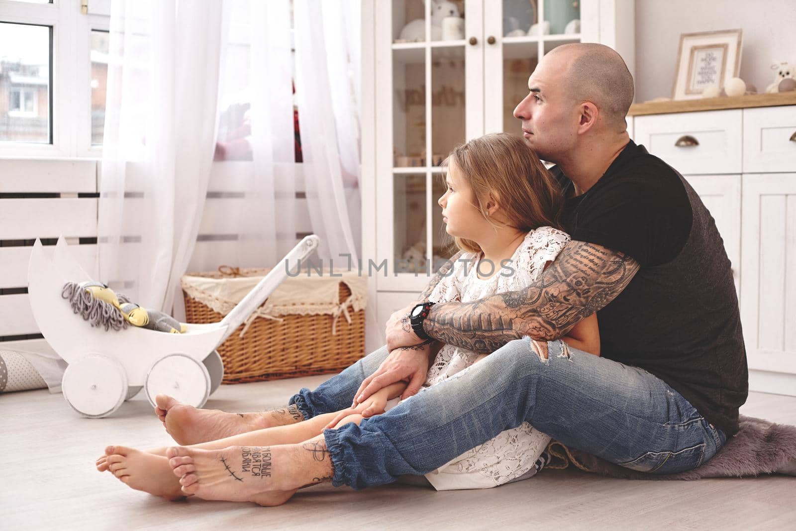 White modern kid's room whith a wooden furniture. Adorable daughter with a long blond hair wearing a white dress. Daddy with tattoos is hugging her and they are looking at the window. Friendly family spending their free time together sitting on a pillows.