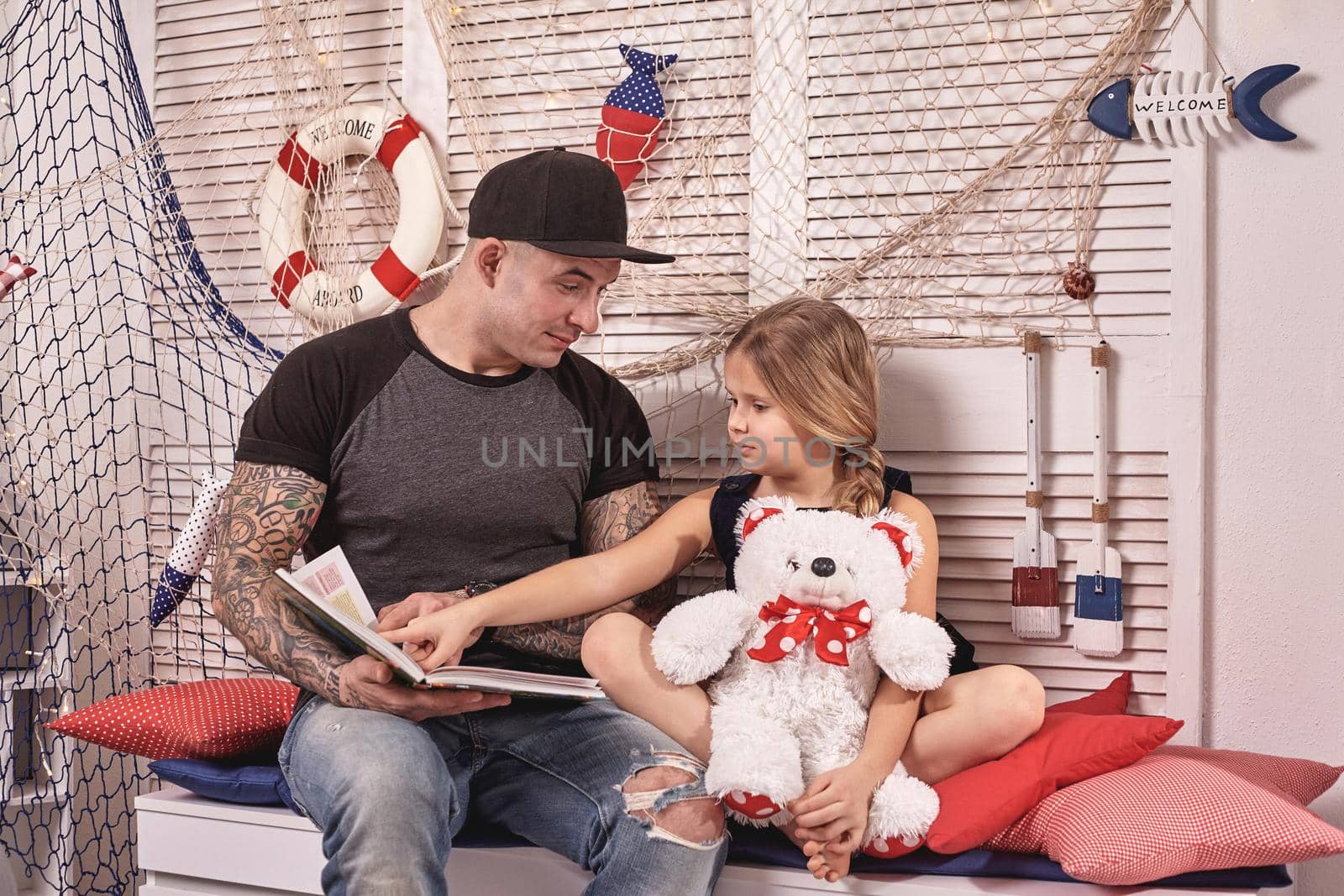 Handsome tattoed man in a cap is spending time with his little cute daughter. They are sitting on a white bench with some red pillows, in a room decorated in a marine style. She is holding a toy bear and pointing on something in a book. Reading fairytales while daughter is sitting nearby. Happy family.