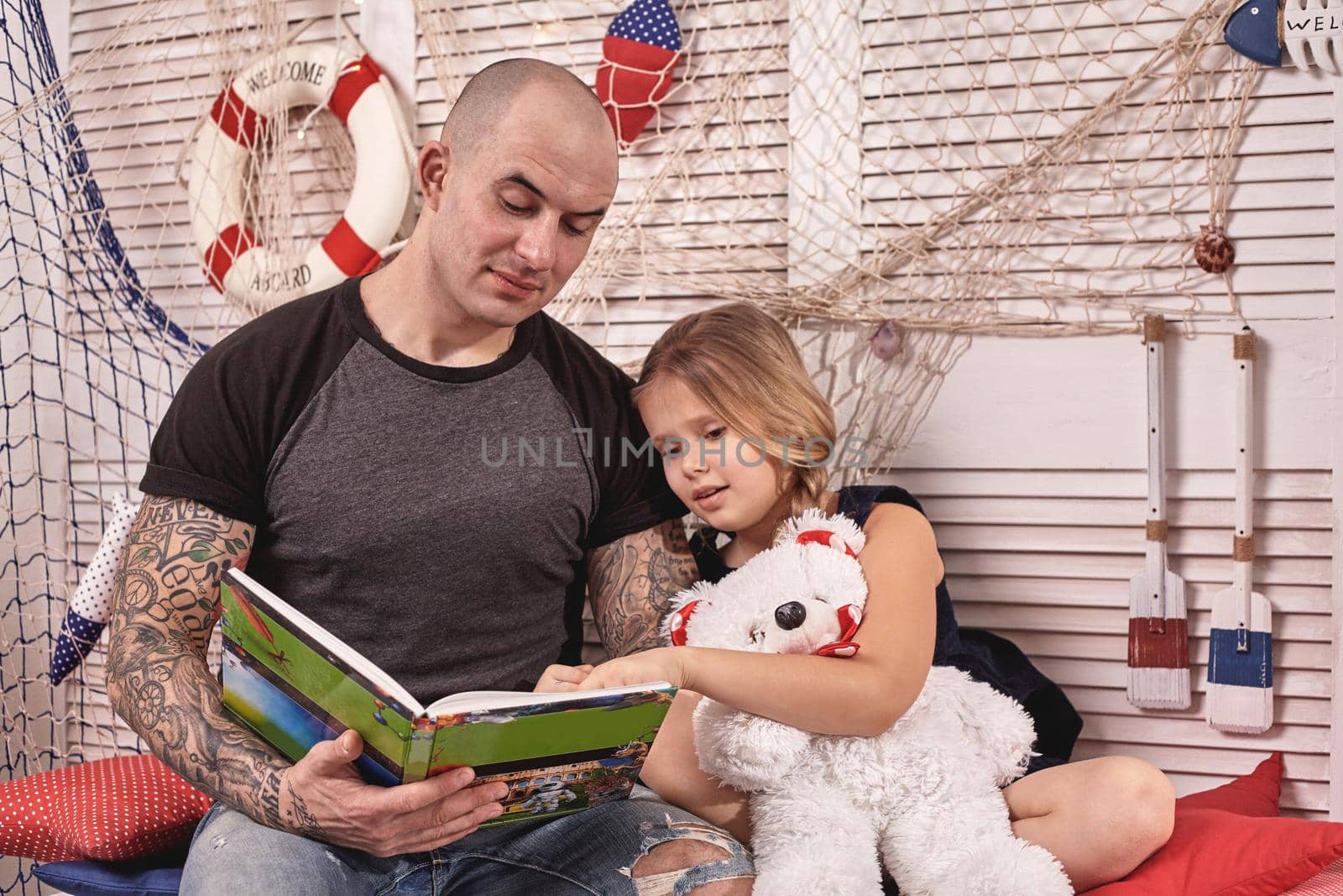 Athletic tattoed man in a cap is spending time with his little cute daughter. They are sitting on a white bench with some red pillows, in a room decorated in a marine style. She is holding a toy bear and looking at the book. Reading fairytales while daughter is sitting nearby. Happy family.