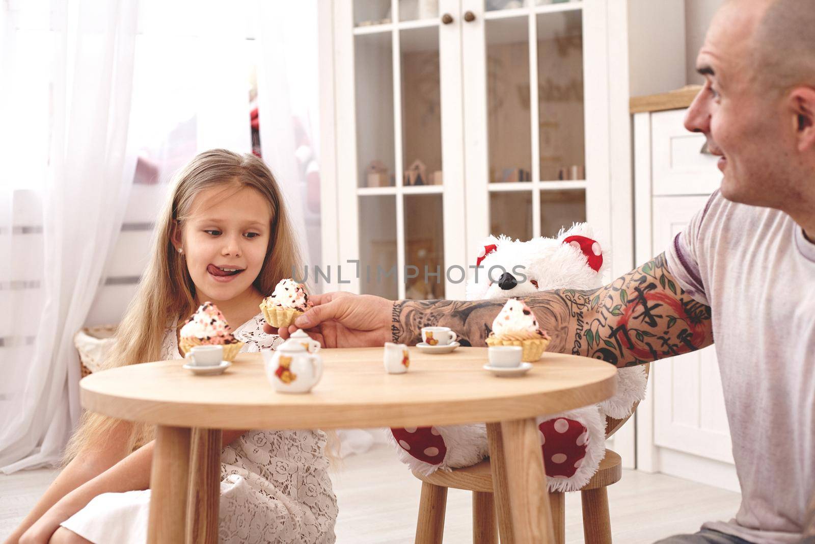 White modern kid's room whith a wooden furniture. Adorable daughter wearing a white dress with a long blond hair. They are drinking tea from a toy dishes in a modern kid's room whith a wooden furniture. Young daddy with tattoos is treating his little daughter with a cupcacke. Friendly family spending their free time together sitting on a pillows.