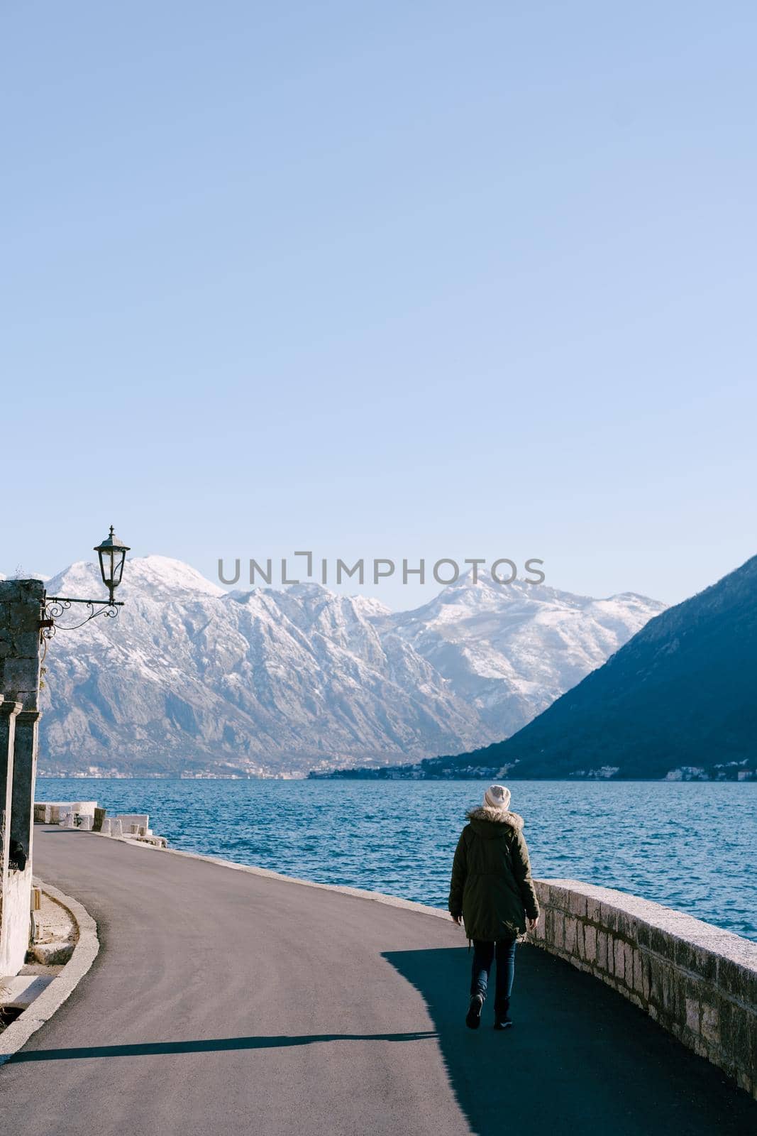 Girl in a jacket walks along the asphalt embankment against the background of the sea and mountains. High quality photo