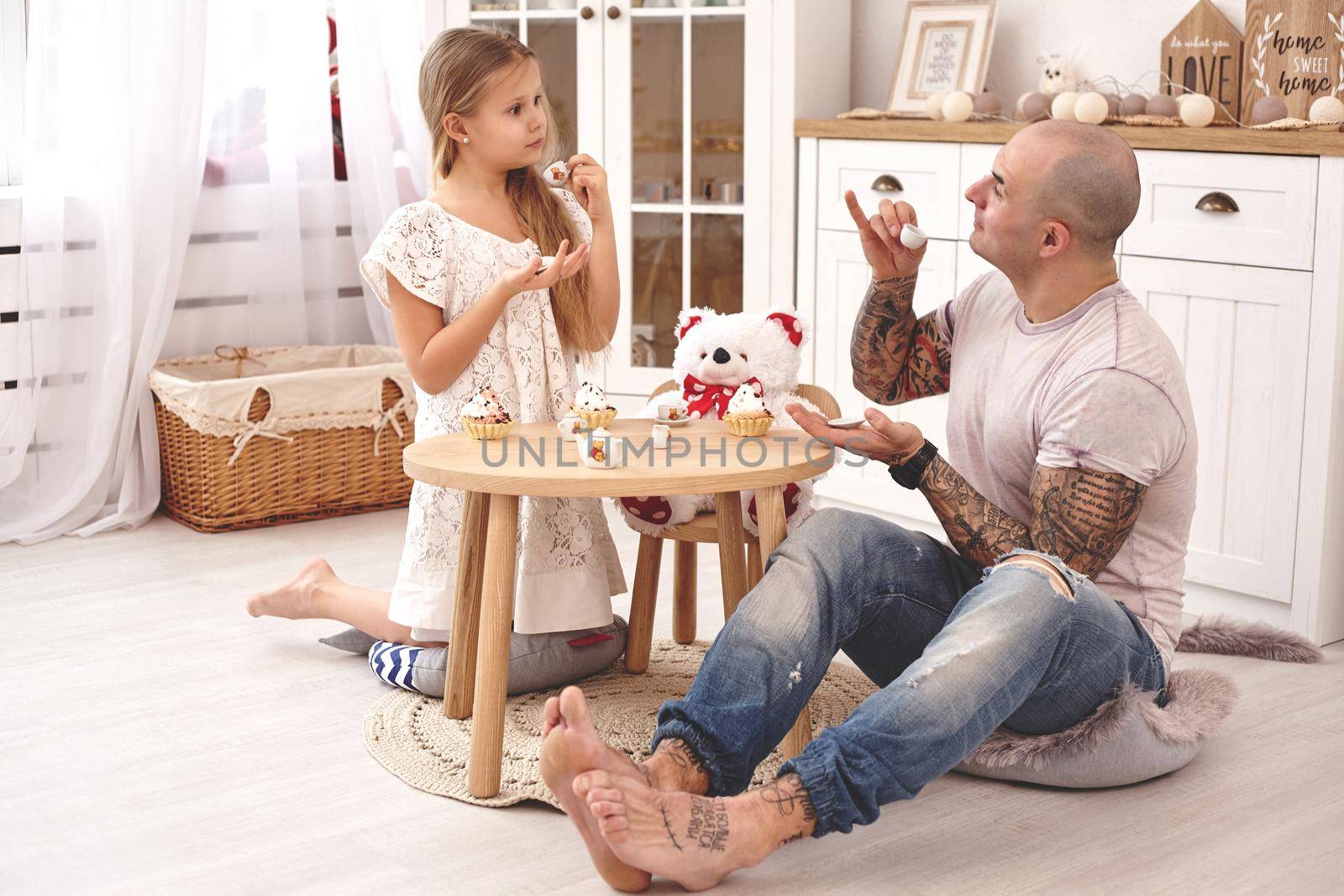 White modern kid's room whith a wooden furniture. Adorable daughter whith a long blond hair wearing a white dress. They are drinking tea with cupcackes from a toy dishes in a modern kid's room whith a wooden furniture. Young daddy with tattoos is looking at his daughter and she is looking at him also. Friendly family spending their free time together sitting on a pillows.