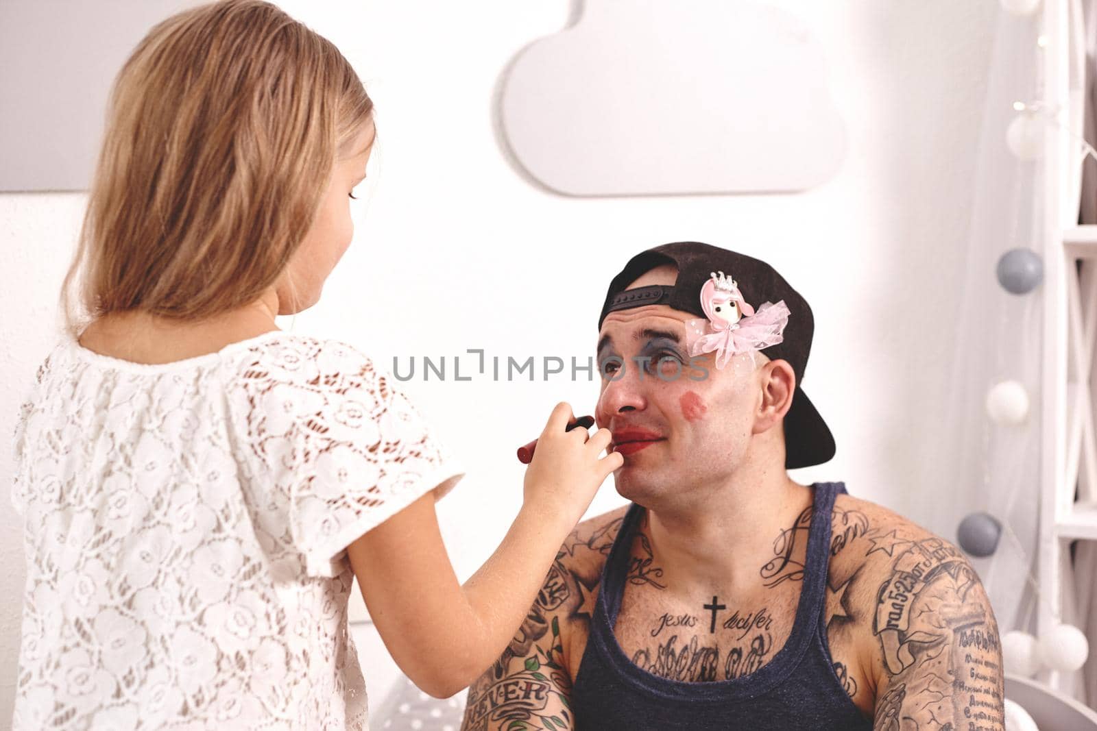 Funny time Tattoed father in a cap and his child are playing at home. Cute girl is doing makeup to her dad in her bedroom. Family holiday togetherness by nazarovsergey