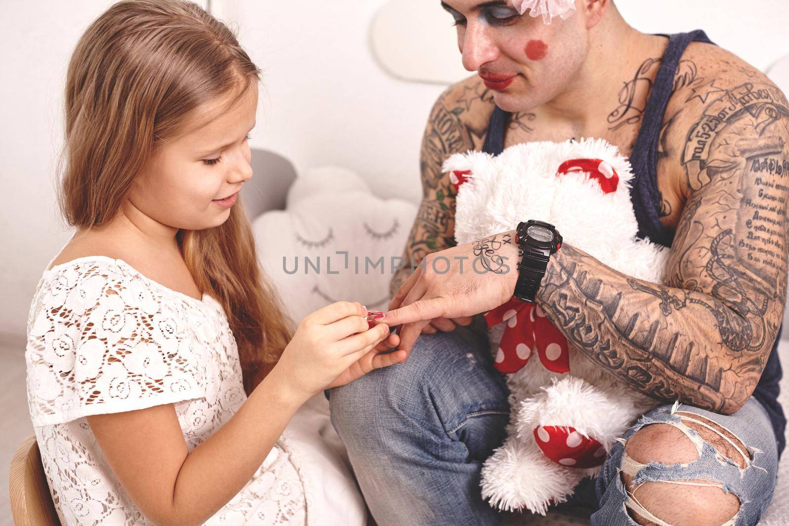 Funny time Tattoed father in a cap, whith a toy bear in his hands, and his child are playing at home. Cute girl in a white dress is applying a red nail polish to her dad's fingers in a bedroom. Family holiday and togetherness.