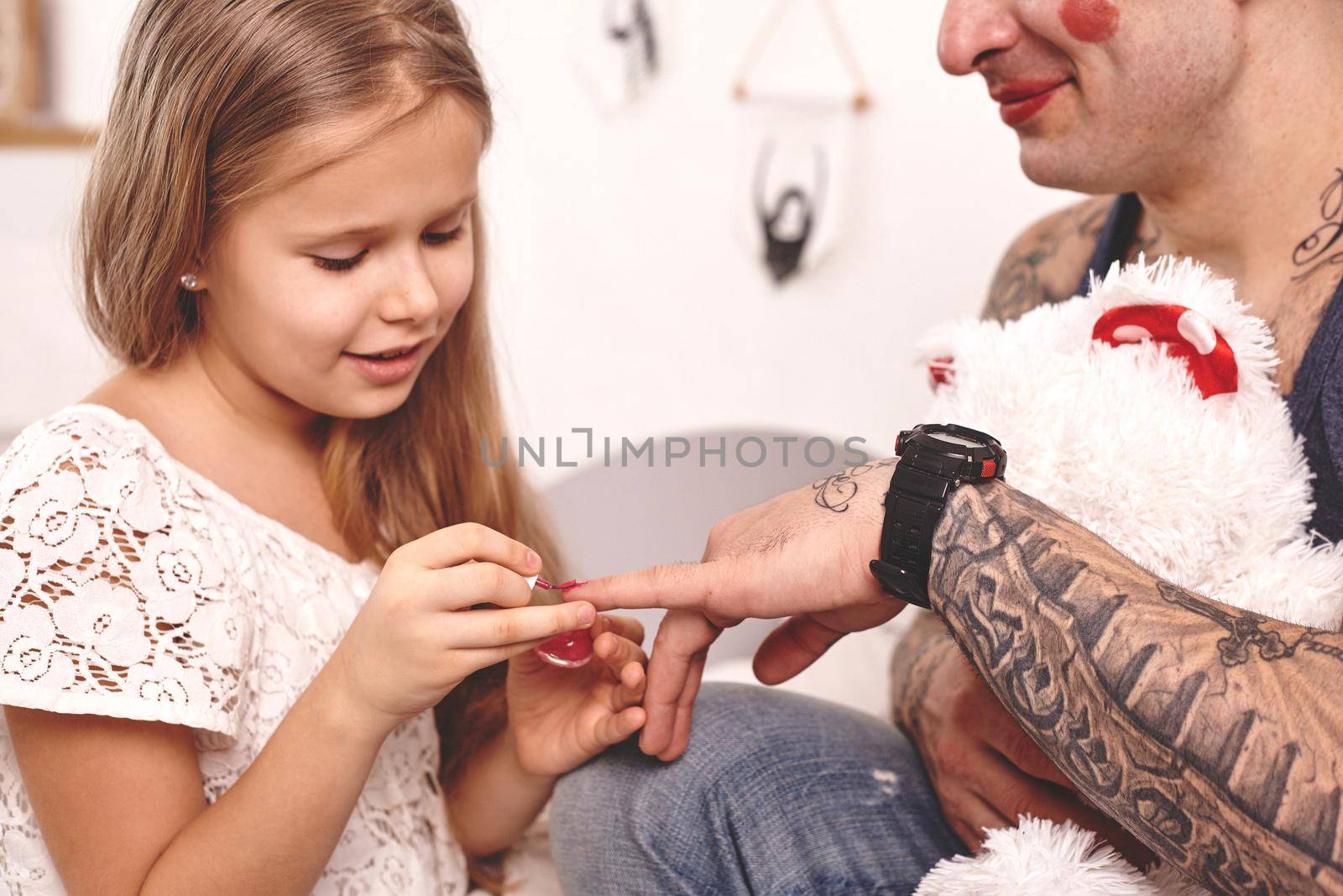 Funny time Tattoed father in a cap, whith a toy bear in his hands, and his child are playing at home. Pretty girl in a white dress is applying a red nail polish to her dad's fingers in a bedroom. Family holiday and togetherness.