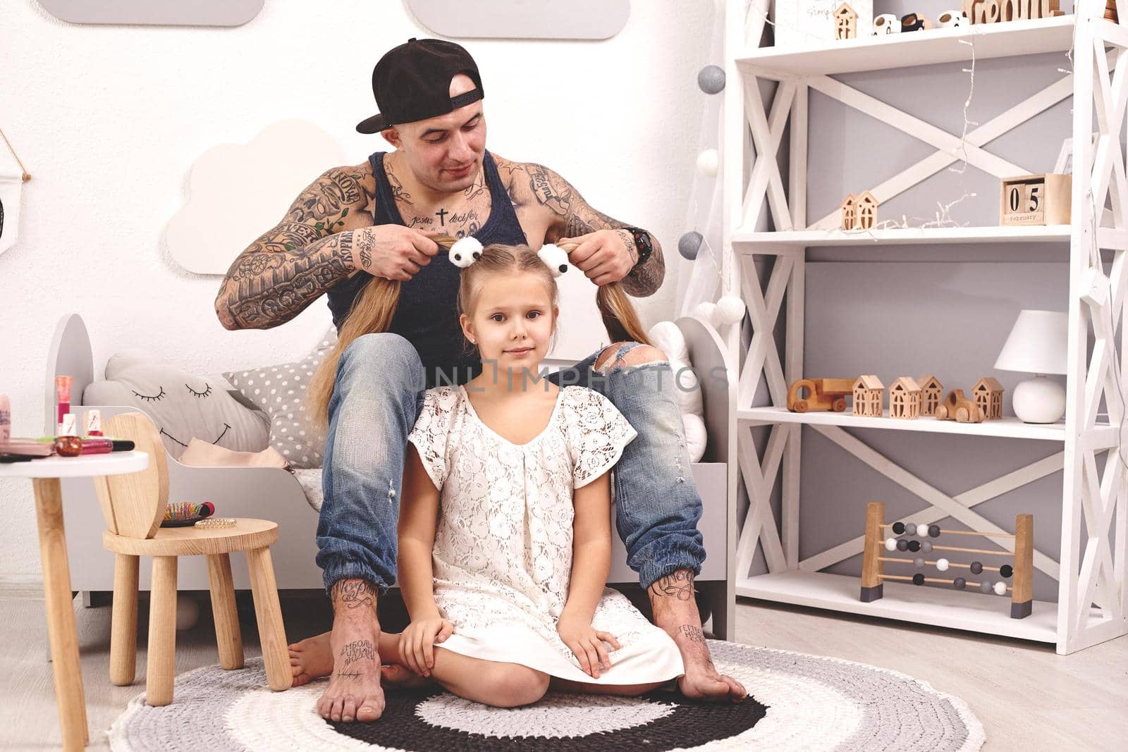 Funny time Tattoed father in a cap and his child are playing at home. Dad is doing his daughter's hair while she is sitting on a rug and smiling in her bedroom. Family holiday and togetherness.