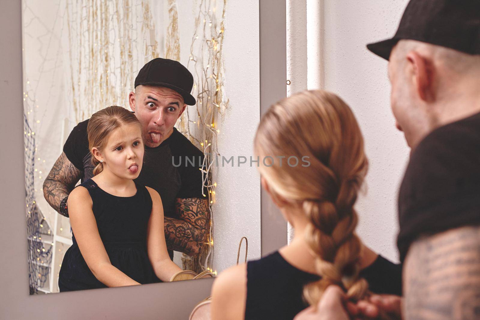 Cute little daughter and her tattoed dad are playing together near a mirror. Dad is doing a braid for his little princess. They are showing tongues to each other. Family holiday and togetherness.