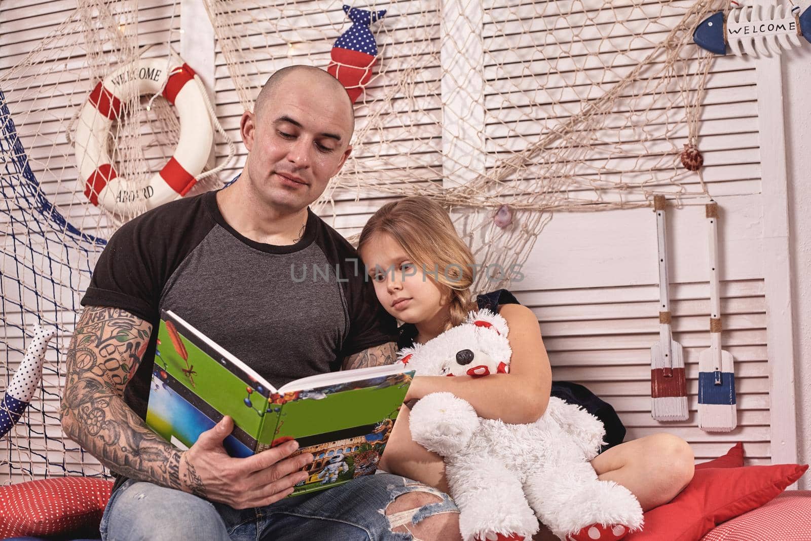 Bald tattoed man in a cap is spending time with his little cute daughter. They are sitting on a white bench with some red pillows, in a room decorated in a marine style. She is holding a toy bear and looking at the book. Reading fairytales while daughter is sitting nearby. Happy family.