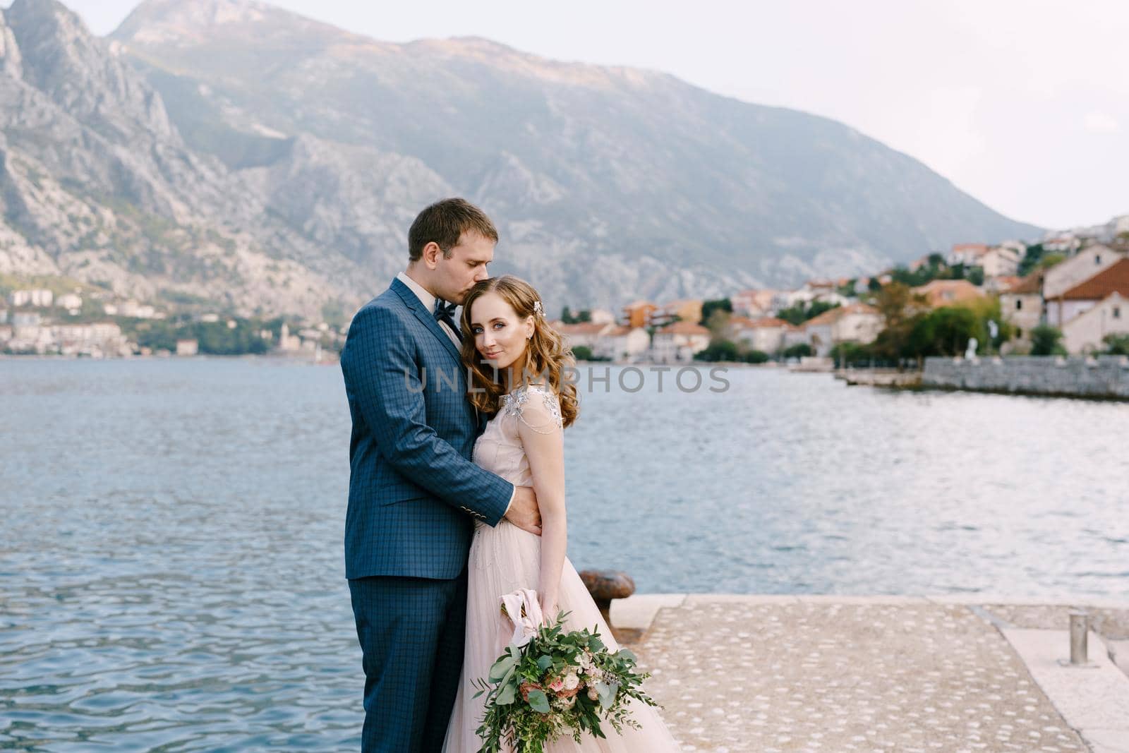 Groom hugs bride on the pier against the backdrop of mountains and the town on the coast. High quality photo