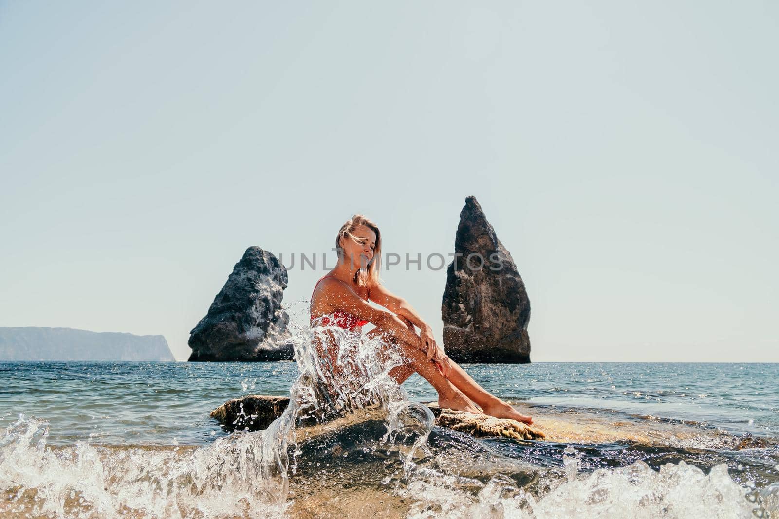 Young sensual blonde woman sitting on the rock near water at sea and enjoys the sea waves on background of two volcanic rocks, like in Iceland. Dreams holidays and weekend vacation in summer time