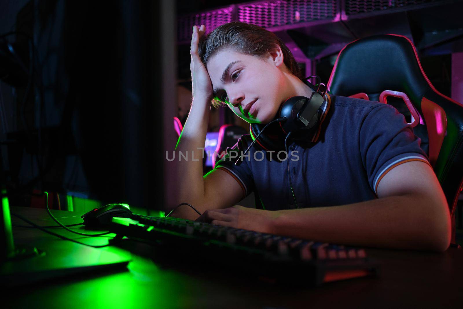 Photo of professional young caucasian gamer playing online on his PC in gaming club or cyber arena. He looks displeased and upset. Game plan went wrong