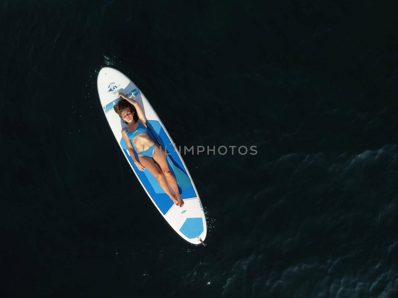 Aerial drone view on young attractive brunette woman with long hair in blue swimsuit, swimming on sup around volcanic rocks, like in Iceland. Summer holiday vacation and travel concept. by panophotograph