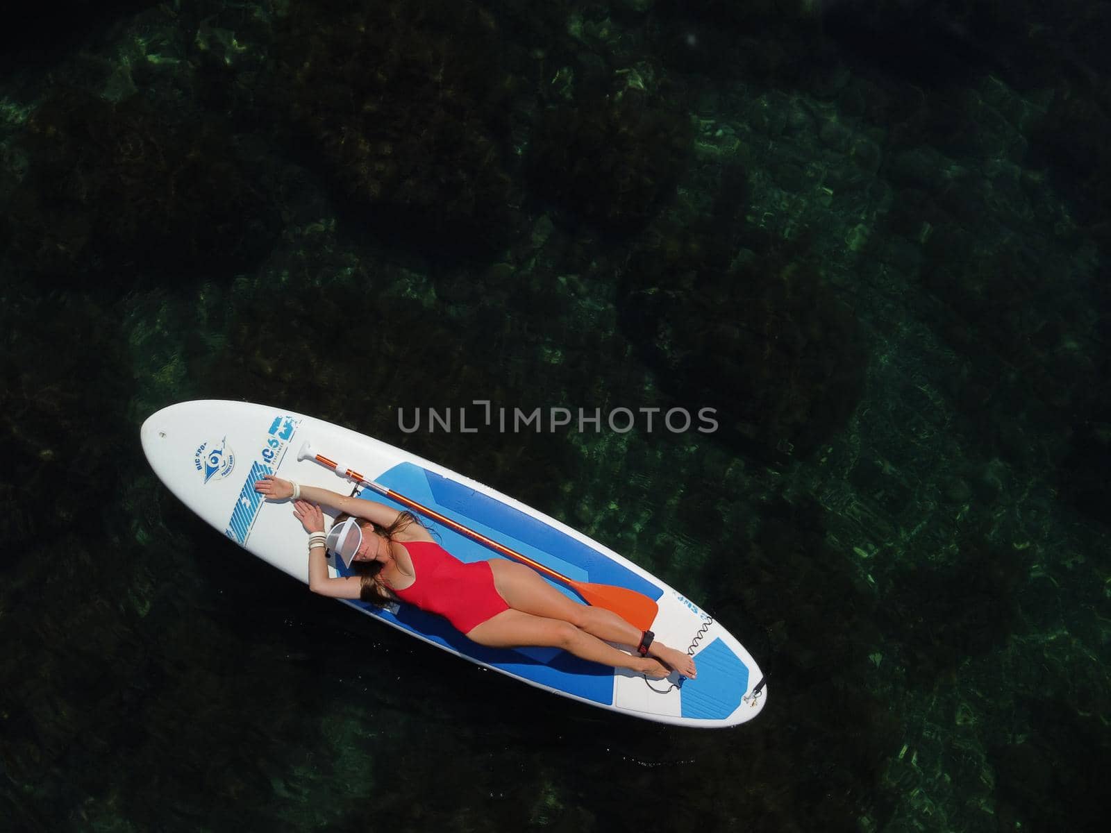 Aerial drone view on young attractive brunette woman with long hair in red swimsuit, swimming on sup around volcanic rocks, like in Iceland. Summer holiday vacation and travel concept. by panophotograph