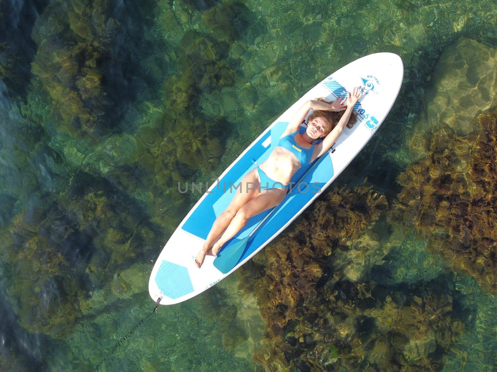 Aerial drone view on young attractive brunette woman with long hair in blue swimsuit, swimming on sup around volcanic rocks, like in Iceland. Summer holiday vacation and travel concept. by panophotograph