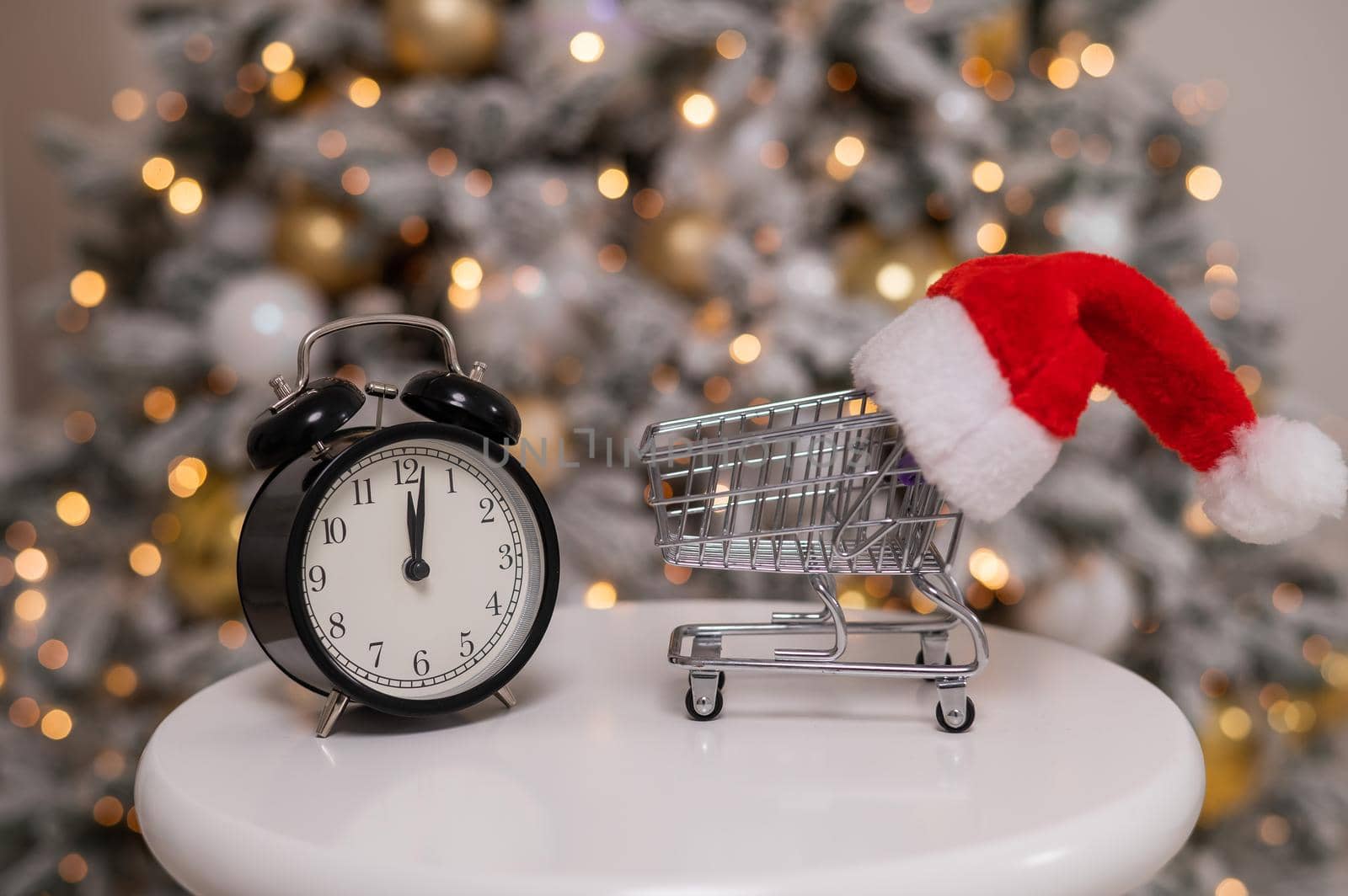 Christmas gifts shopping time. A shopping trolley with a santa hat and an alarm clock by the Christmas tree