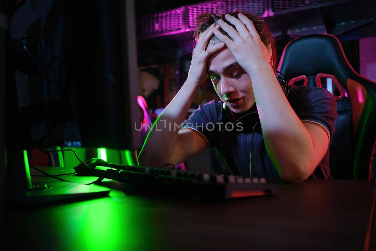 Photo of professional young caucasian gamer playing online on his PC in gaming club or cyber arena. He looks displeased and upset. Game plan went wrong