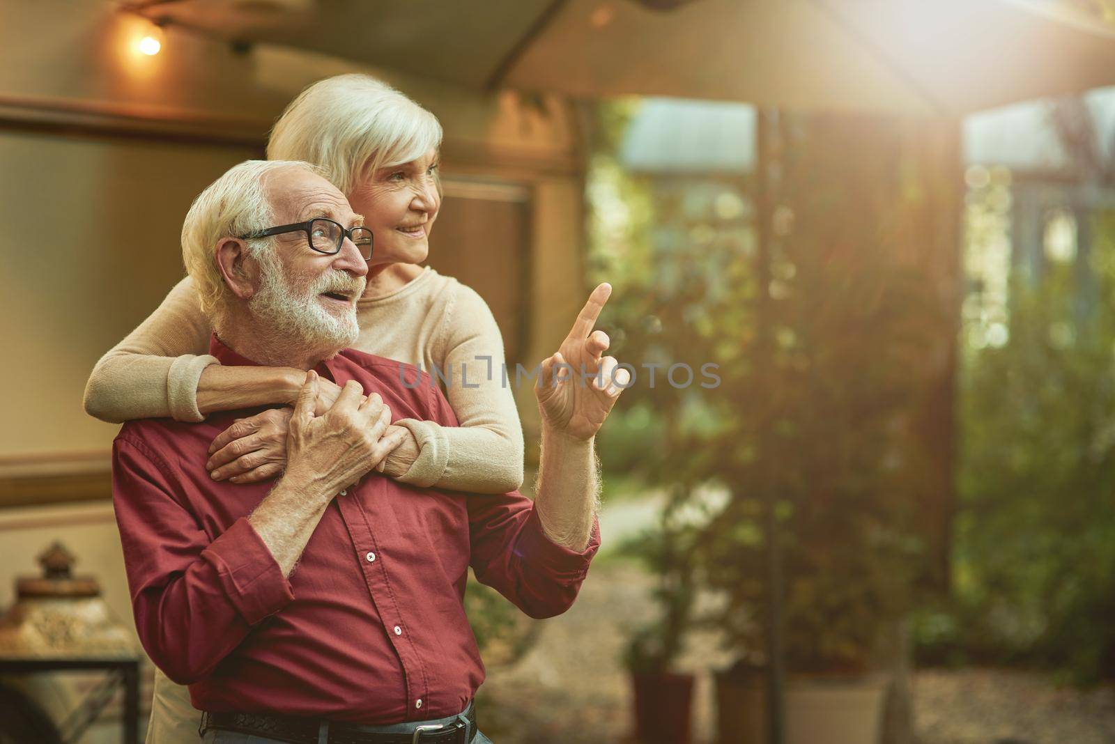 Smiling male retiree in glasses pointing finger to the side and embracing with his wife by friendsstock