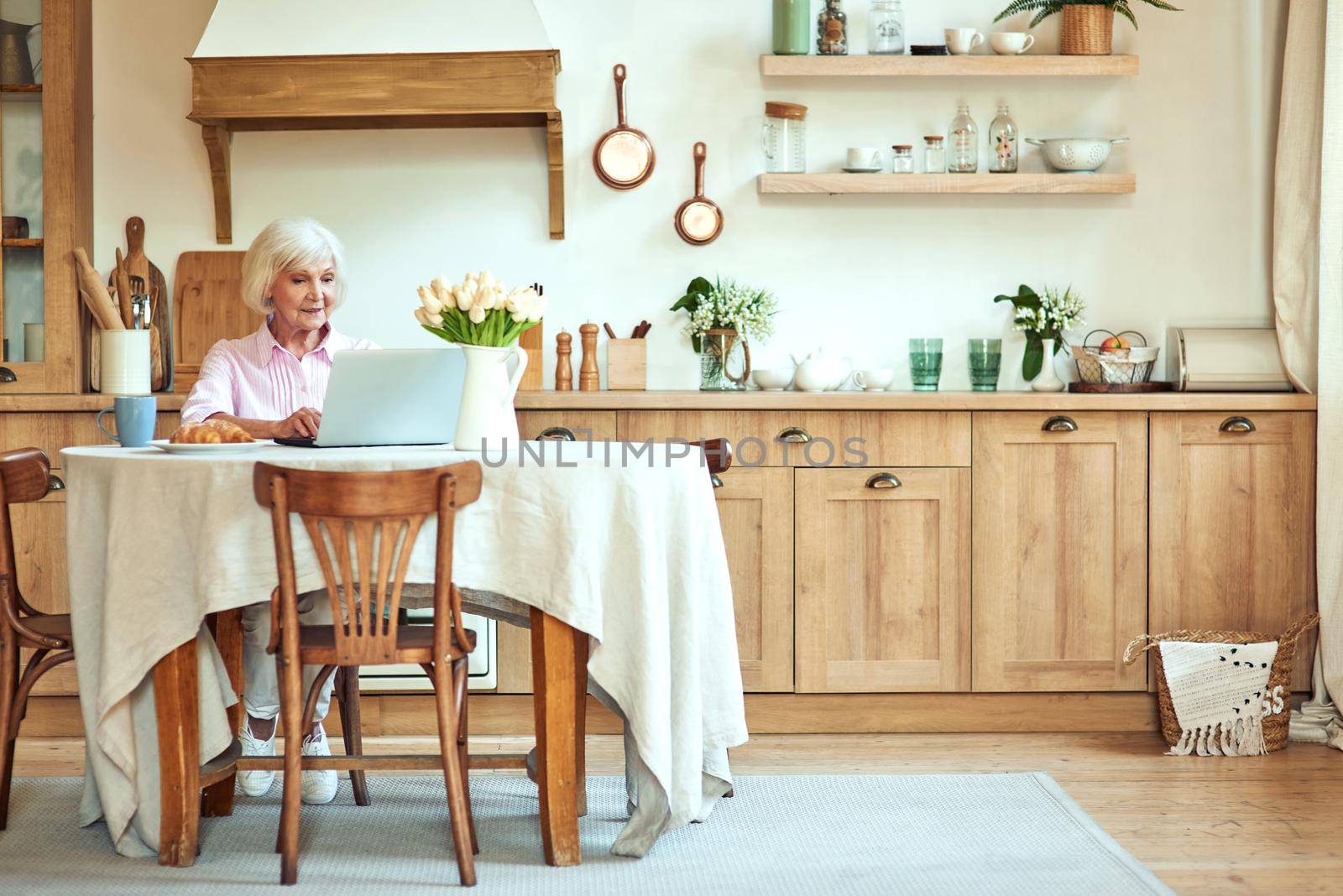 Elderly lady typing on laptop while sitting at the table at home during morning. Domestic lifestyle concept. Copy space