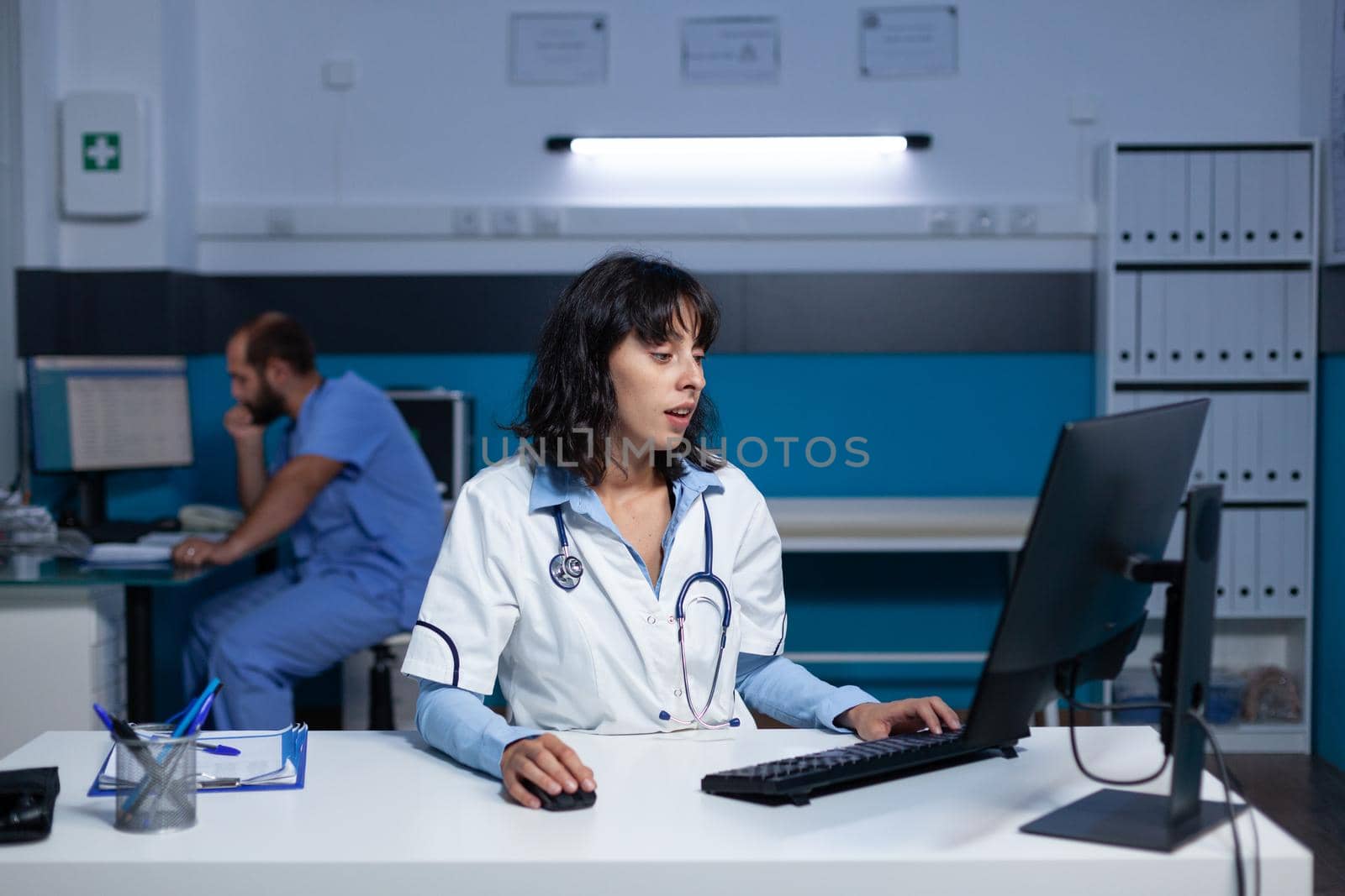 Medical physician typing on computer keyboard at office, working late at night. Woman doctor with stethoscope looking at monitor display for healthcare system and assistance after hours