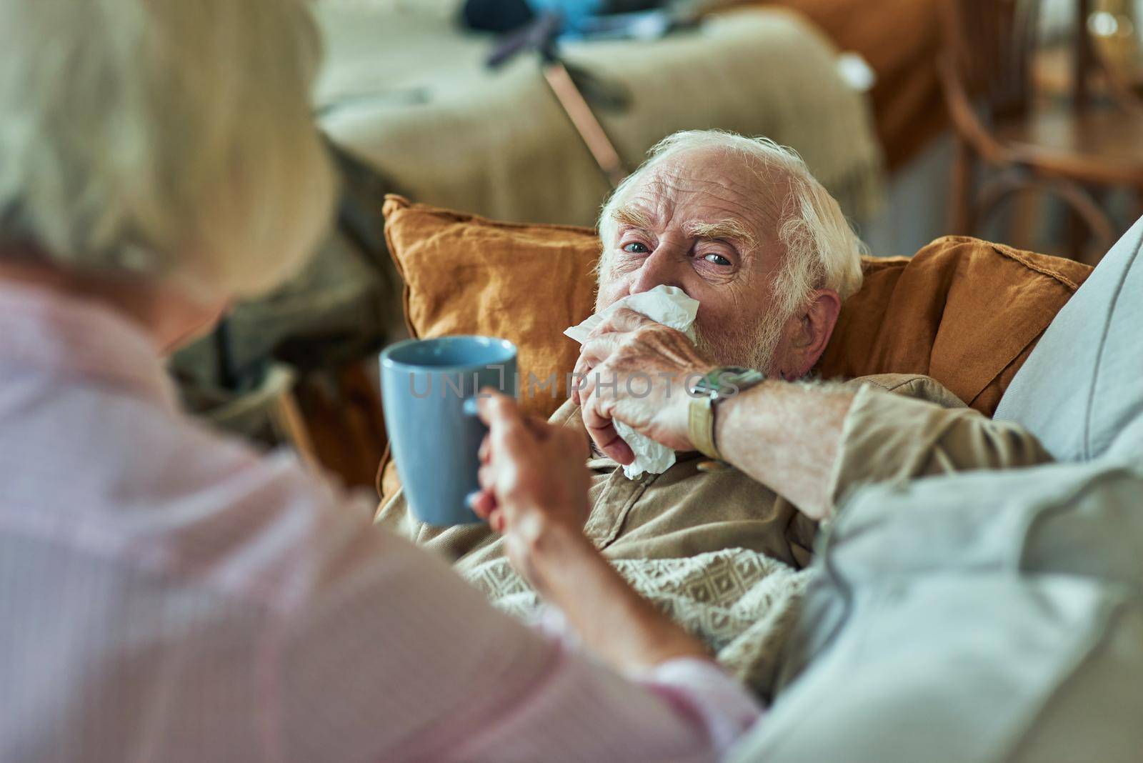 Sick elderly man lying on the couch and using his napkin for a runny nose with wife next to him. Care and health concept