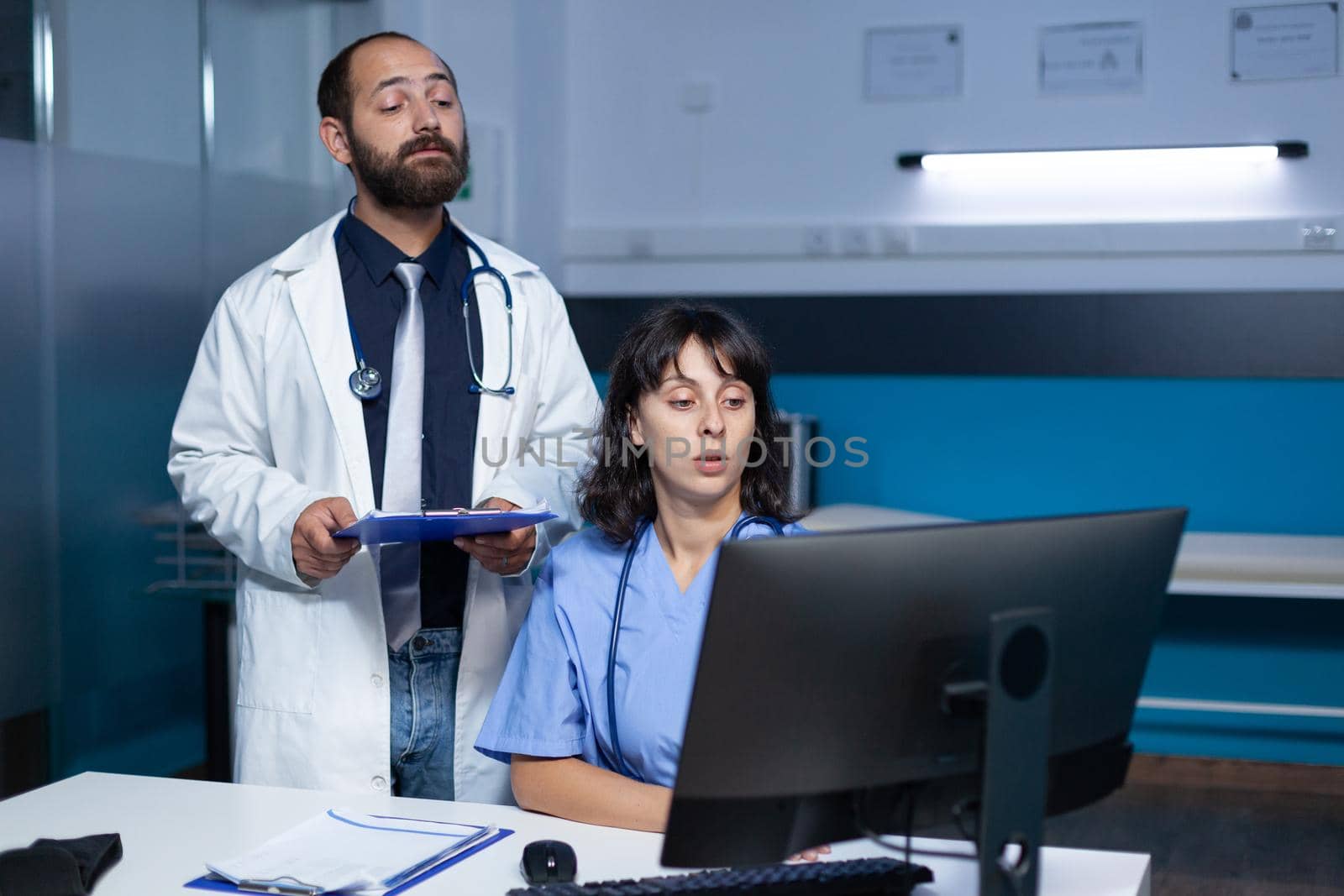 Doctor and nurse working with technology on computer for medical care and treatment. Team of workers using monitor and checkup files for healthcare system, working late at night.