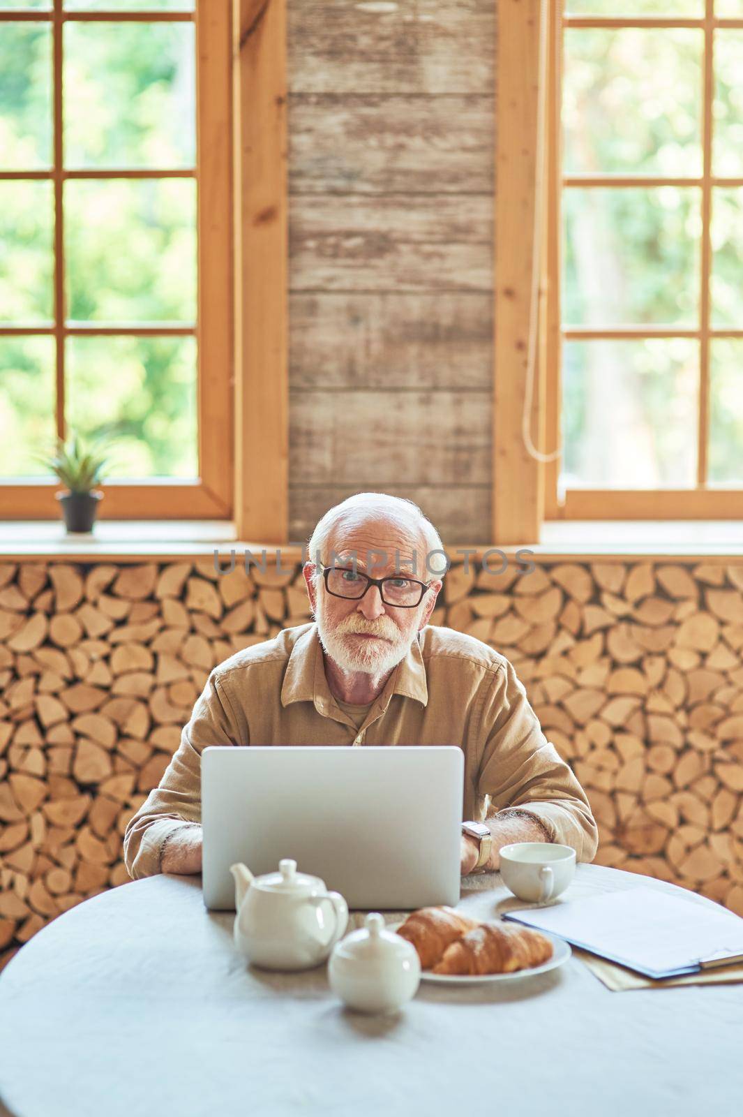 Serious elderly man sitting in room at home while using laptop and having breakfast. Lifestyle concept