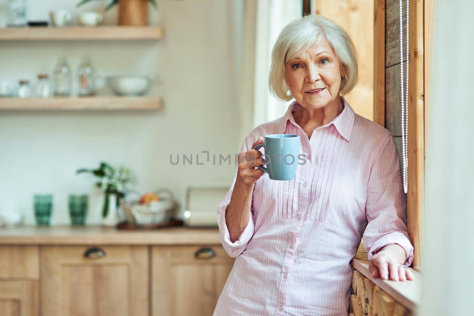 Waist up of smiling stylish gray haired lady standing near window and holding cup of coffee at the kitchen. Domestic lifestyle concept
