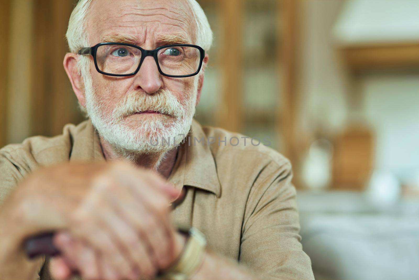 Close up of concentrated male pensioner in glasses resting at home while looking away and holding hands on walking stick. Care and health concept