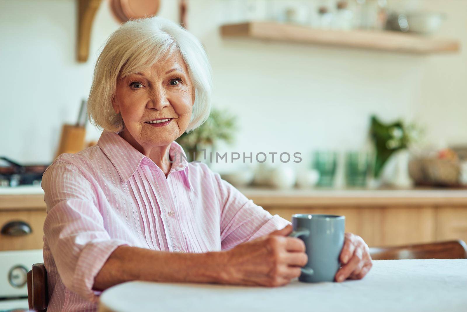 Waist up of happy beautiful senior lady sitting at the kitchen in her cozy house while enjoying hot drink. Domestic lifestyle concept