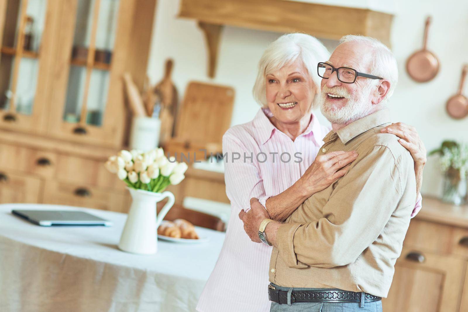 Waist up of happy senior man and woman hugging while posing at camera at their kitchen. Lifestyle concept