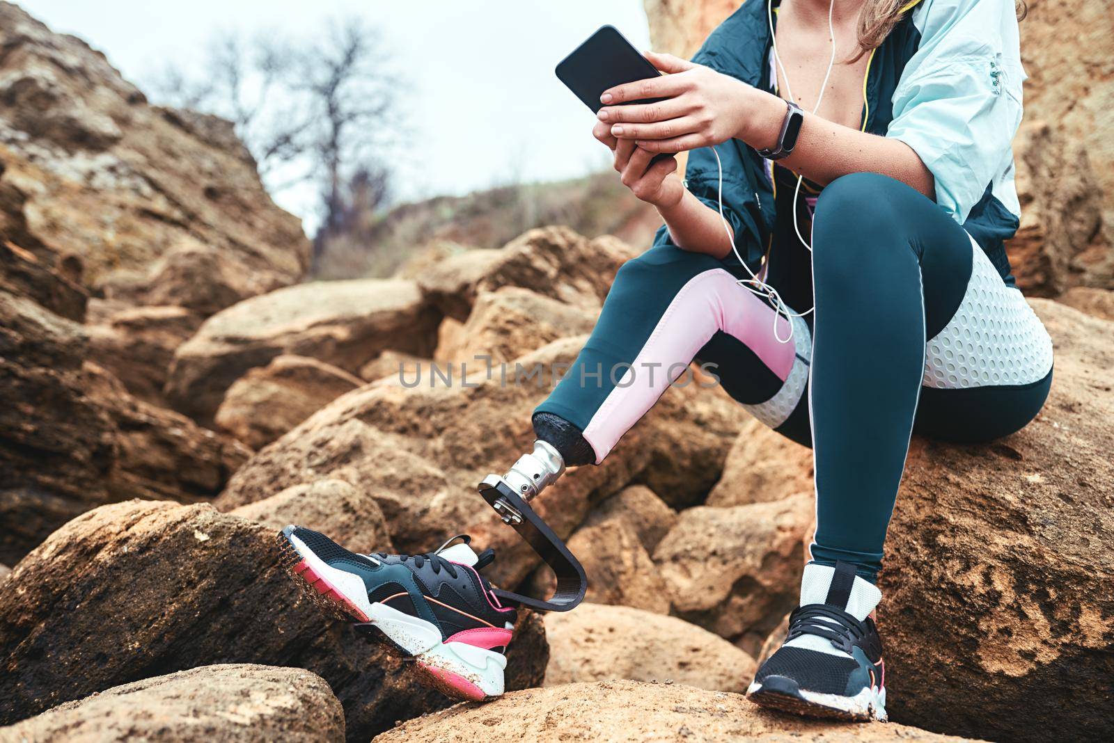 Digital technologies. Cropped image of disabled women in sport wear with leg prosthesis sitting on the boulders and holding smartphone. by friendsstock