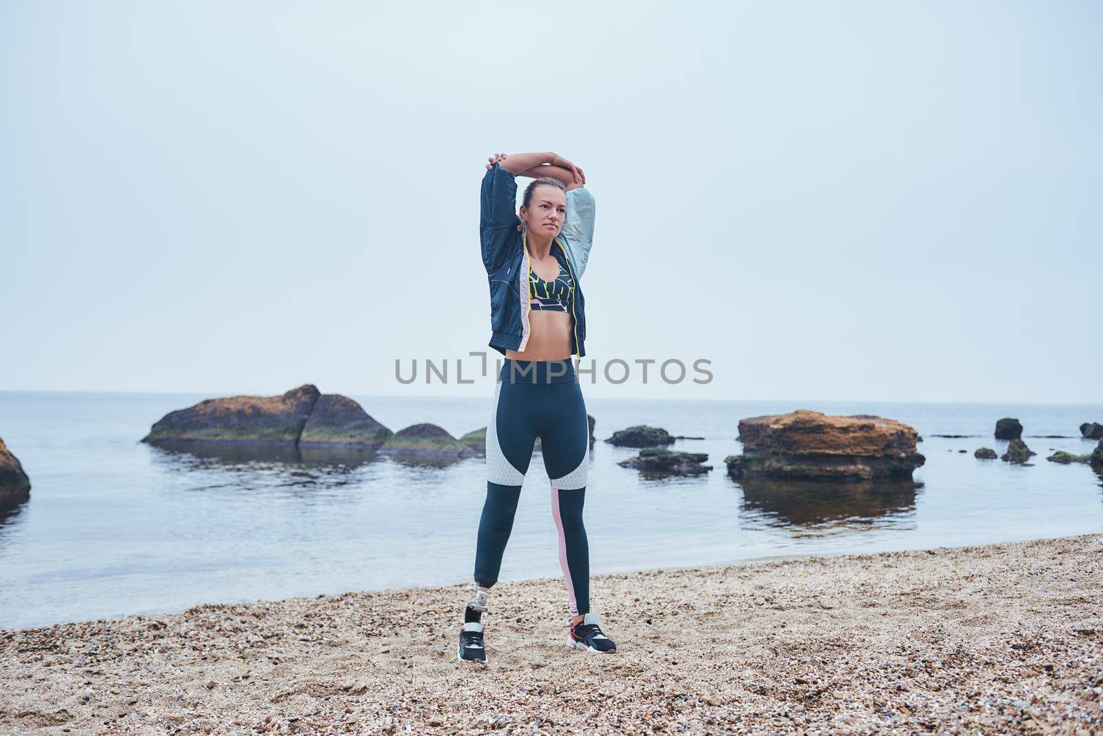 Breathe deep. Strong disabled athlete woman with prosthetic leg doing stretching exercises while standing at the beach by friendsstock