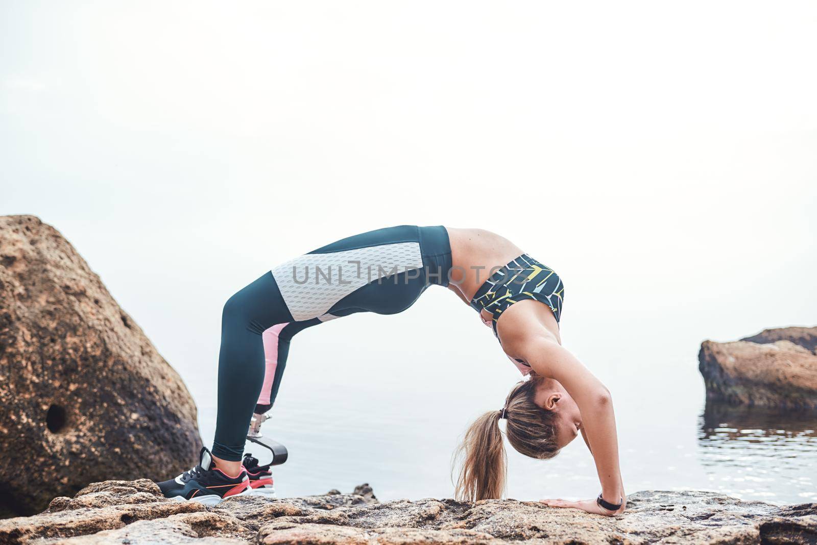 Flexible body. Young disabled athlete woman in sportswear with prosthetic leg making a gymnastic exercise bridge on the the stone in front of the beach by friendsstock