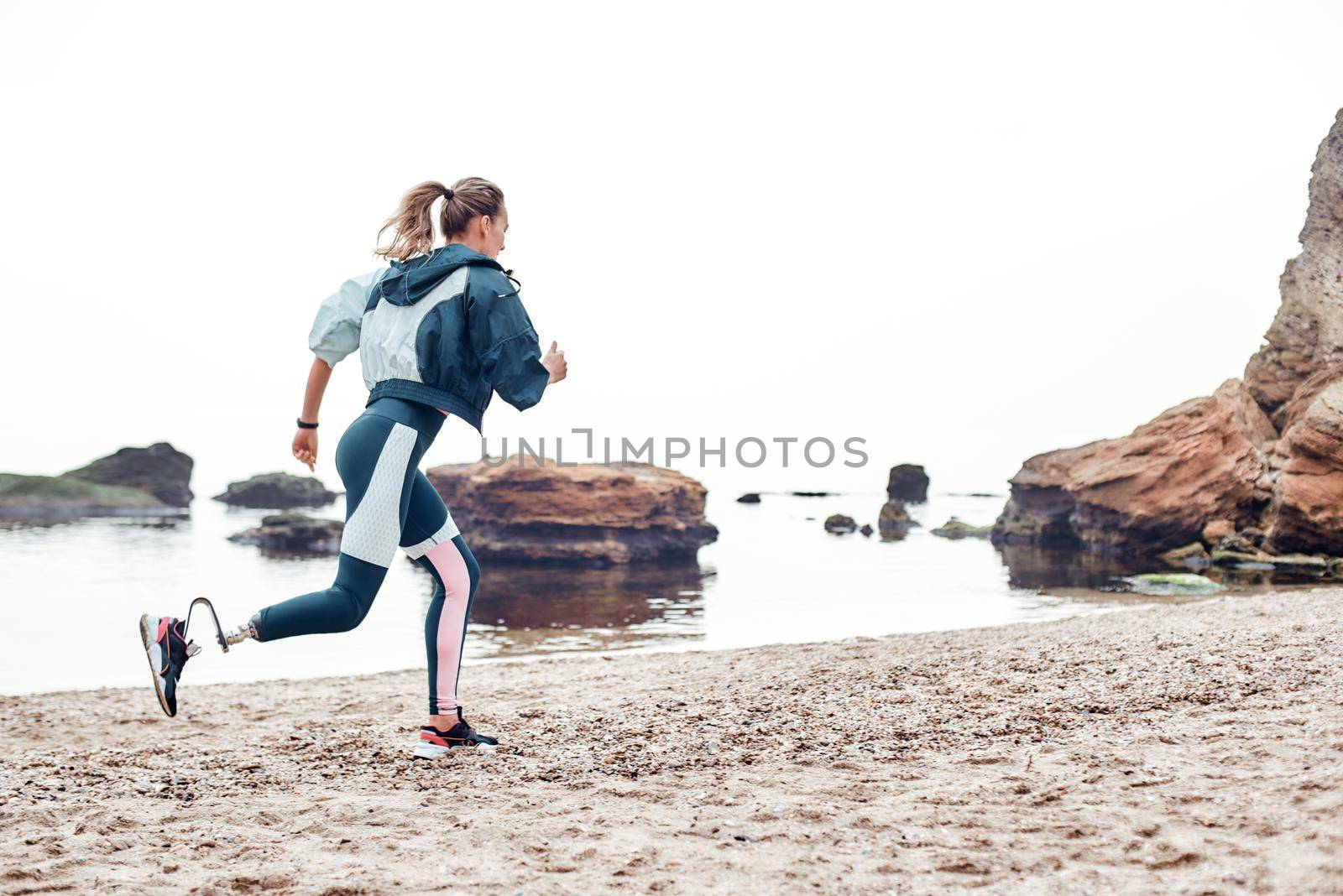 Take care of your body. Side view of strong disabled woman in sportswear with prosthetic leg is running on the beach. Motivation. Sport concept. Disabled Sportsman