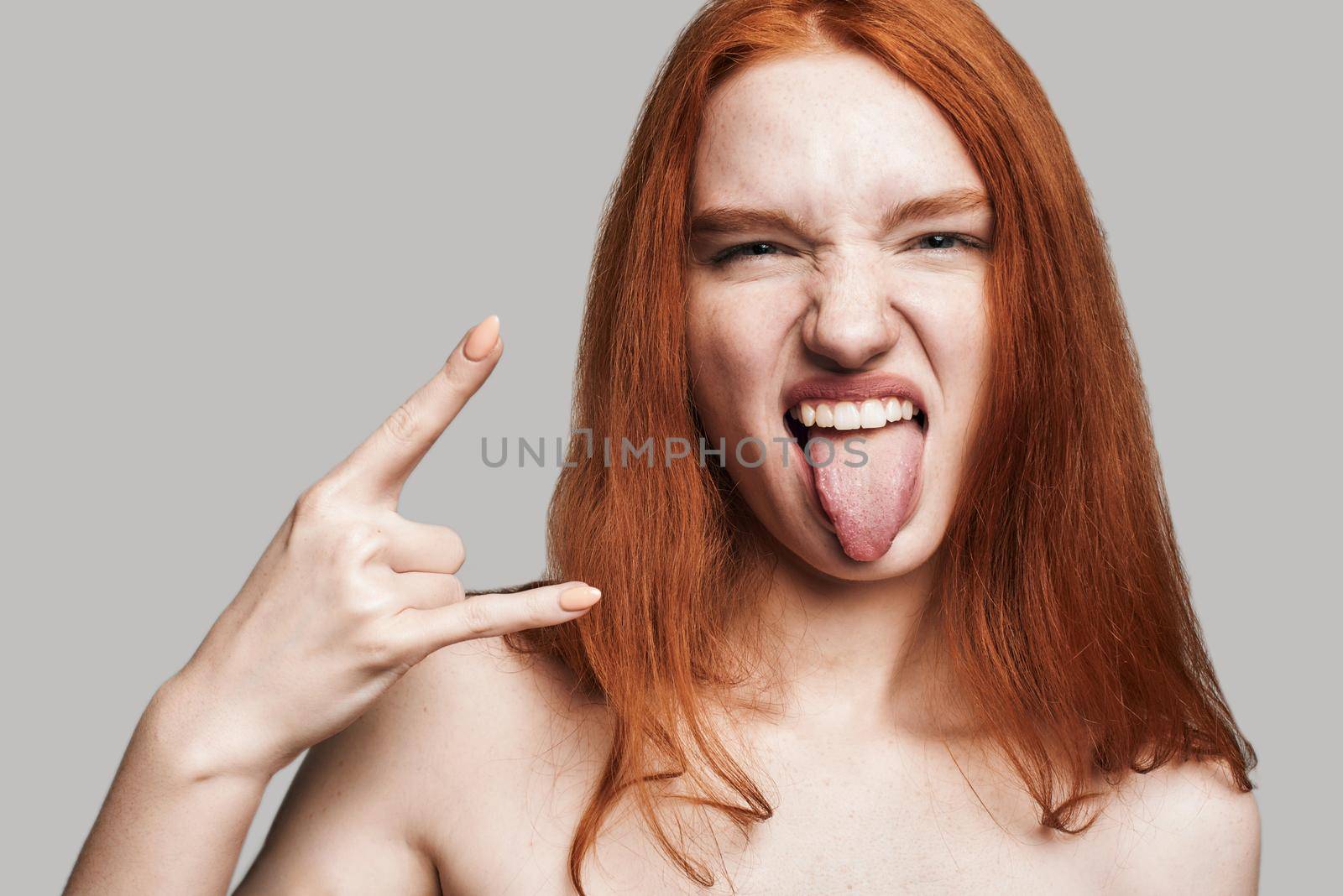 Studio shot of young and cute teenage girl with long red hair showing hand sign and sticking out tongue while standing against grey background. Gesturing. Carefree