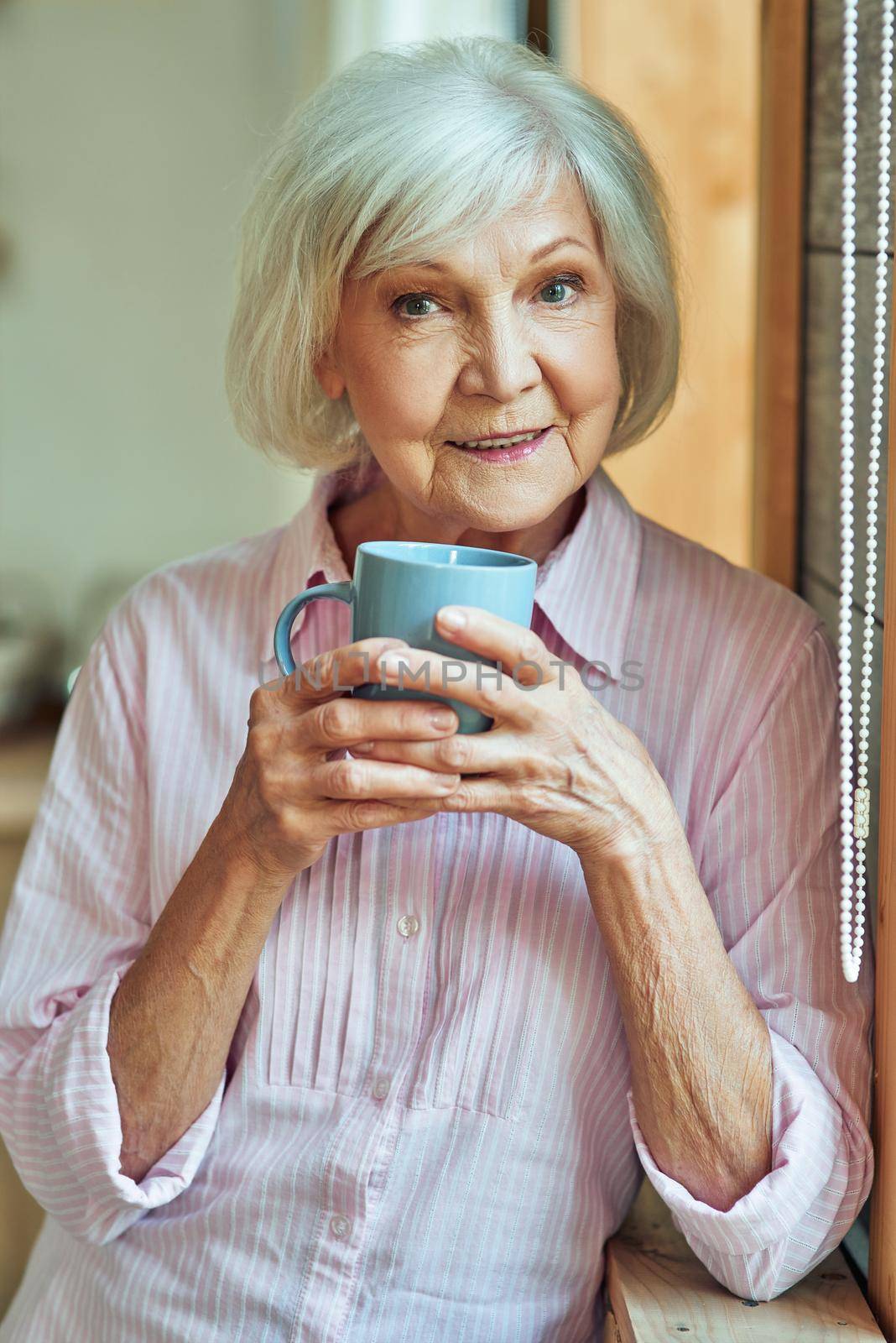Happy stylish gray haired lady standing near window with cup by friendsstock