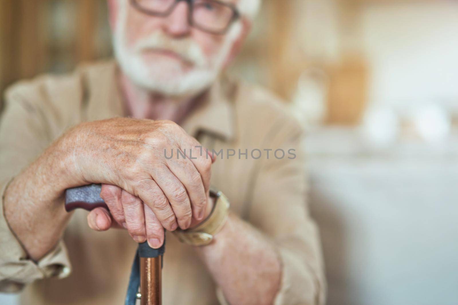 Cropped photo of senior bearded man resting at home and holding wooden walking stick. Focus on cane. Care and health concept
