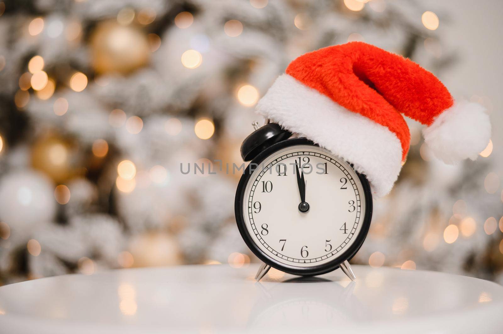 Alarm clock in a santa hat against the background of a christmas tree