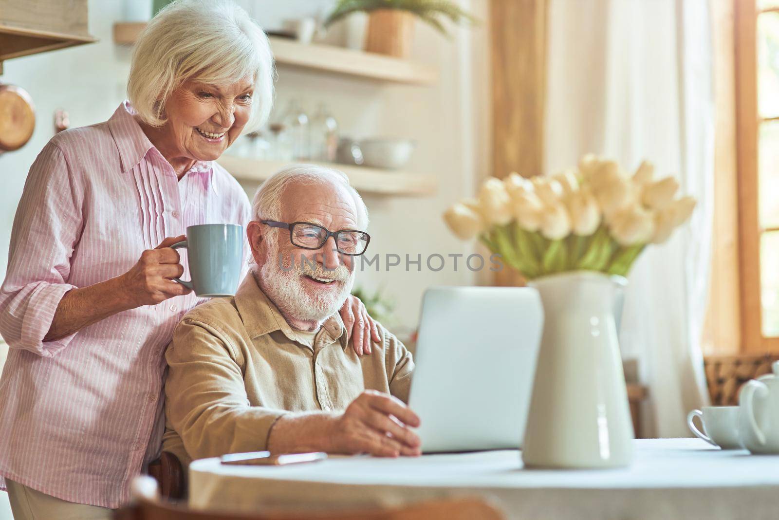 Happy elderly couple using laptop while spending morning together at the kitchen by friendsstock