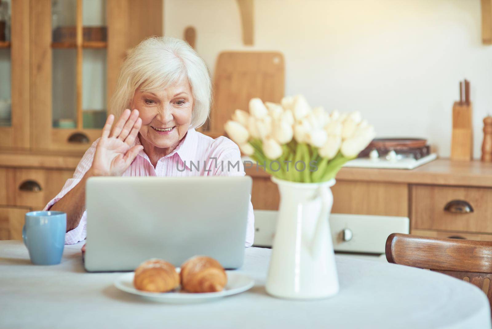 Happy elderly lady sitting at the table and making video call on laptop in kitchen at home. Domestic lifestyle concept