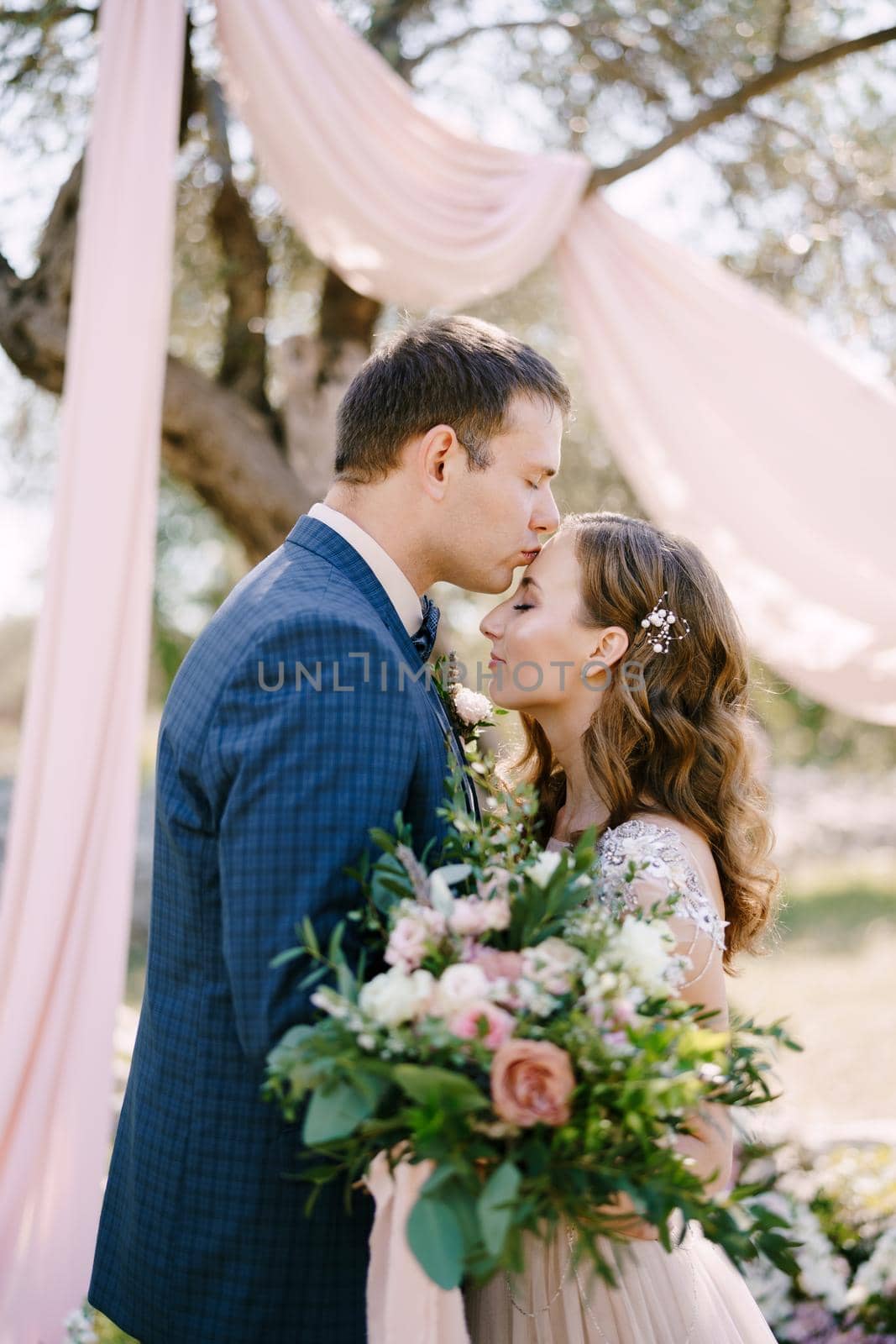 Groom kisses bride on the forehead with a bouquet of flowers under a tree. Portrait by Nadtochiy